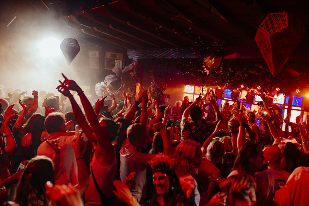 Crowd of participants dancing with hands raised during a The Gardens of Babylon event, featuring immersive red lighting and an atmosphere reflecting the collective’s narrative and spiritual approach