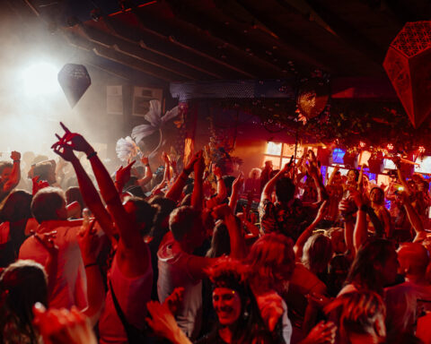 Crowd of participants dancing with hands raised during a The Gardens of Babylon event, featuring immersive red lighting and an atmosphere reflecting the collective’s narrative and spiritual approach